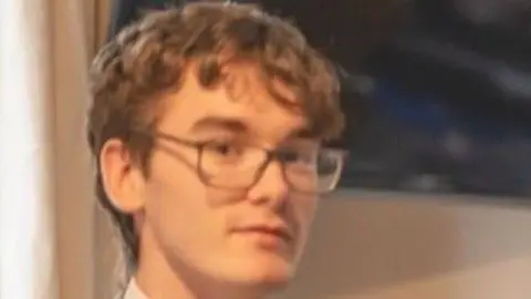 A teenage with short brown hair and glasses looks at the camera