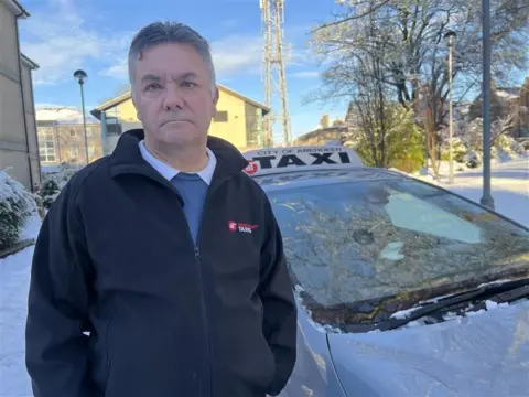 Stuart Benzie, who has grey hair, is standing next to his taxi. The ground around him, in a residential area, is covered in snow. 
