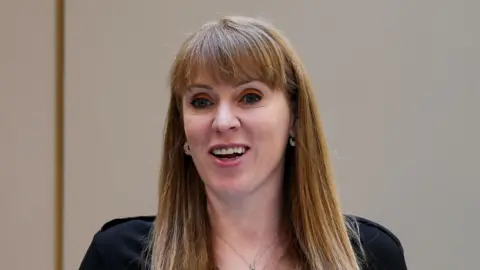 Angela Rayner smiles while looking beyond the camera. She has long red hair with a fringe. She wears a black top as well as earrings and a necklace.