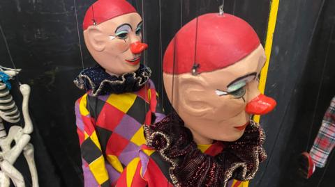 Sad looking clown marionettes in black, red, yellow and purple-checked smocks. They have red paint on their noses and red hair nets