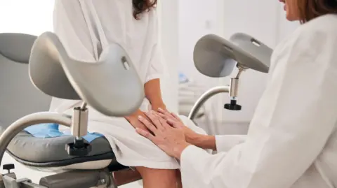 The young woman in a hospital gown sits on the stretcher as the mature doctor in a white lab coat, seated on a stool, holds her hands.