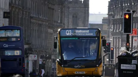 The Q3 which is a single-decker yellow and black bus. The board on the front reads 'Wallsend via City Centre & Quayside'. The bus has stopped at a red light on a street lined by stone buildings.