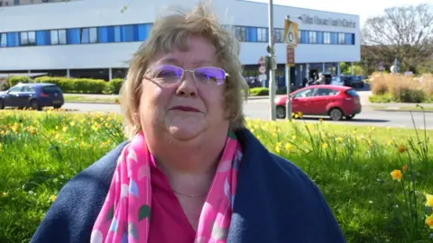 A woman with collar length fair hair and glasses looking at the camera. She is wearing a pink top and scarf with colourful large dots on it, and a blue coat. She is surrounded by grass with yellow daffodils. A road is behind her with a couple of cars in the picture.