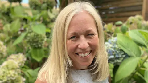 Cathy Neville has blonde shoulder-length hair and is smiling at the camera. Behind her are some blue and pink hydrangeas in front of a wooden fence.