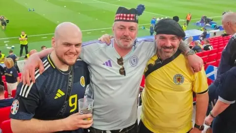 Robert McCahill Robert McCahill stands between his brother Stephen, on the right, and fellow Scotland fan Darren McConway. They are all inside a football stadium and are wearing Scotland tops.