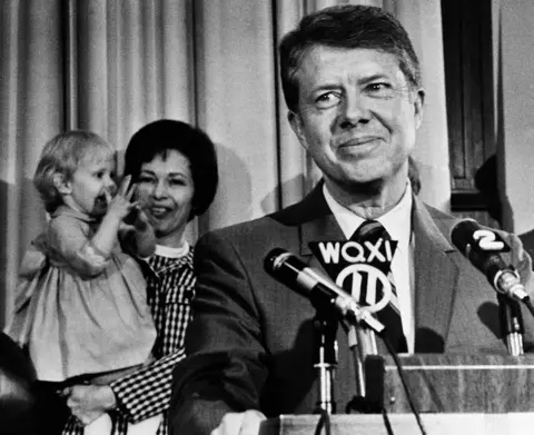 Ish-senatori i AP Jimmy Carter dëgjon duartrokitje në Kapitol në Atlanta më 3 prill 1970, pasi shpalli kandidaturën ose guvernatorin e tij. Në sfond, gruaja e tij Rosalyn mban vajzën dyvjeçare Amy, e cila u bashkua me duartrokitjet.