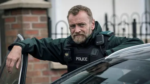 MILK A man wears a green top with a black police stab vest over it. He has short brown hair and a short grey and brown beard. His right hand leans on an open car door, whilst his left hand leans on the roof of the car. 