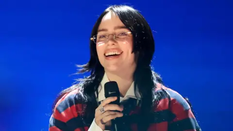 A close up head and shoulders shot of Billie Eilish smiling and singing into a microphone while performing at the FireAid show in Inglewood, California on 30 January