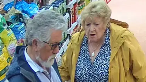 Two elderly people pictured on a CCTV image inside a garden centre. The man on the left has grey hair and a grey beard and wears sunglasses. The woman has short blond hair and a blue and white patterned blouse, and a yellow anorak.