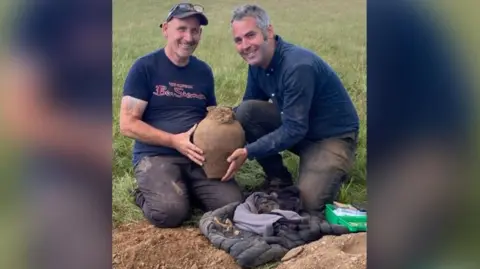 Minelab Two men holding a clay pot smiling with the dig site seen just in front of them. The man on the left has glasses on his head and a cap, he has a blue t-shirt with the words Ben Sherman, in red font, written on the front. The man on the right has salt and pepper hair, a blue shirt and blue pants. 