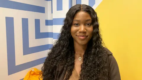 A woman with long curly black hair stands by a yellow painted wall looking at the camera. 