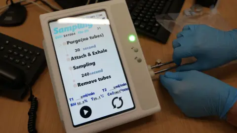 A small white unit with a screen sitting on a desk. Its screen reads: sampling, purge, attach and exhale and remove tubes, and there are also some readings including for Co2. A pair of hands in blue latex gloves is visible next to the machine attaching pipes to the side. 