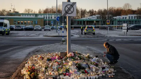 Joel Gunter/BBC A cluster of candles and flowers lies on the frosty ground as a man crouches in front of the scene