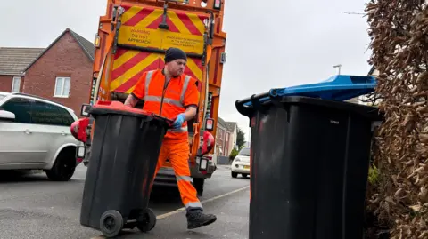 Ben Schofield/BBC A refuse collector walking a recycling bin back to the pavement. In the foreground is a blue-lidded black bin, next to a bush. The bin collector is wheeling an orange-lidded bin from the road and onto the pavement. He is wearing an orange, high-visibility suit, protective gloves and a black beanie-type hat. Behind him is the back of a bin lorry, stationary on the road, between cars. There are houses in the very background. The sky is overcast. 