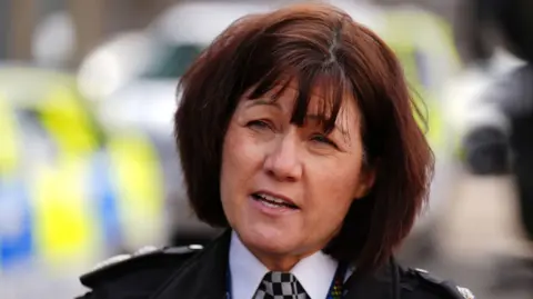 Jo Farrell has short dark brown hair and is mid-speech. She stands in front of a blurred police vehicle and wears a black jacket, white shirt, and black and white checked police tie