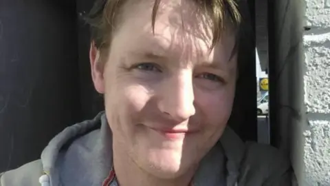 Jamie Forbes leaning against a white wall, smiling at the camera. He is wearing a grey hooded jumper. 