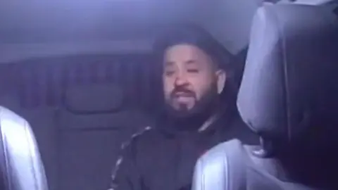 A man sitting in the back of a police car in a black hoodie. He has a black beard and short black hair