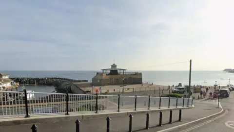 Google Ventnor beach and harbour wall - it is a calm, sunny day. Cars are parked along the road and people can be seen walking to and from the beach.
