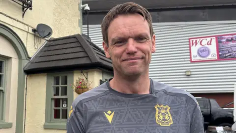 Wayne Jones smiling at the camera. He is stood outside the Turf Hotel which stands next to the Racecourse stadium. he has a grey Wrexham shirt on with gold embroidery. He has short, dark hair. Behind is the pub and the Wrexham football ground.