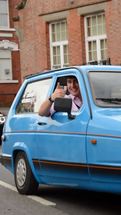 Person smiling and giving a thumbs‑up while sitting in a blue vintage car.