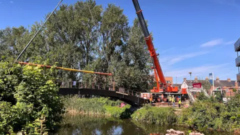 A crane lifting a bridge out of place over a river. It's a sunny day and there are green trees and blue sky. Bushes and grass flank the river.