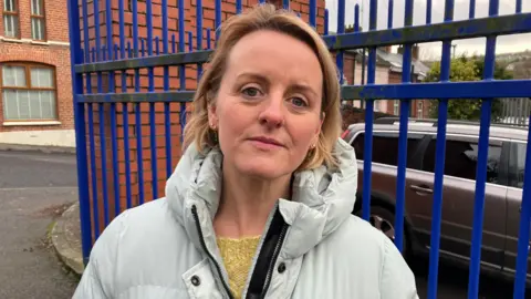 BBC Róisín Blackery stands outside Nazareth House Primary School in Derry. She is wearing a light grey winter coat, and yellow jumper. She is standing in front of a blue gate and the red brick building can be seen behind her