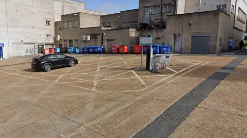 Google Maps The Marks & Spencer Rear Car Park has one black car parked in it. It is lined with industrial bins and the backs of shops. There is a row of shopping trolleys parked next to the car park sign.