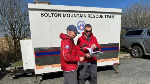 Two men in red jackets pour over a map beneath a white trailer bearing the words 'Bolton Mountain Rescue Team'. They are stood in a car park. 