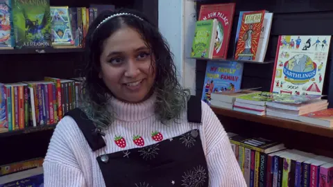 A woman with black curly hair died light green at the end, wearing black dungarees and a corduroy white turtle neck jumper underneath with three strawberries on it. She is sat in the corner of a bookshop with shelved books behind her.