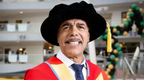 KM IMAGES/UNIVERSITY OF BRADFORD Chris Kamara wears a graduation gown and hat, which has a yellow tassle dropping down. He wears a white shirt and blue tie underneath the gown.