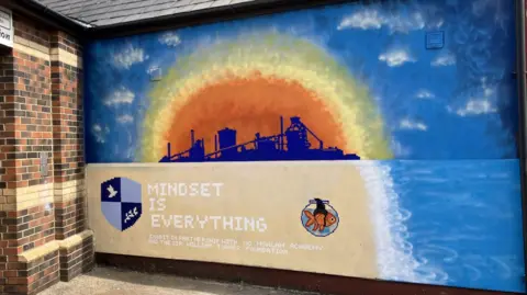 Peter Harris/BBC The artwork shows the former factory in silhouette within an orange sun. It is surrounded by blue sky.