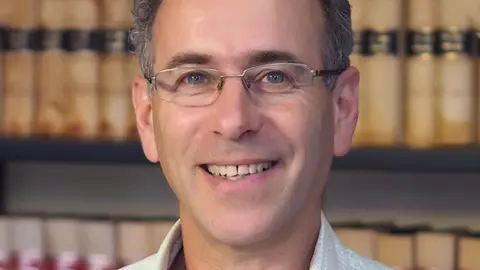 Roland Browne Roland Browne smiles looking directly at the camera. He has grey short hair and is wearing wire-framed glasses. There are books on a shelf in the background.