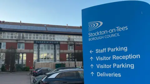 At the right-hand side of the photo is a blue sign outside the Stockton-on-Tees Borough Council building. On it are directions for 'Staff Parking', 'Visitor Reception', 'Visitor Parking' and 'Deliveries'. In the background, part of the car park is visible, and so is the 3-storey building. A sign is at the front of the building that says 'Dunedin House'. The building is made up on predominantly red brick, with many windows and also solar panels on the slanted roof.