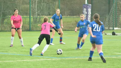 Poucher PR Five girls playing football on a pitch. Three girls are dressed in blue kits and two are in red and pink kits. 