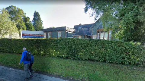 Google A Google Street View screengrab of the Keswick Urgent Treatment Centre. The shot is taken from the street and there are two people walking on the kerb next to the white, red and blue sign for the hospital. There is a grass verge and low hedge around the hospital, which is a slate-clad building.