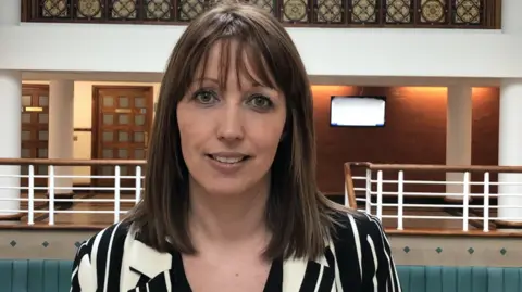 Sarah, with brown hair and fringe, wears a black-and-white shirt in an official-looking public building.