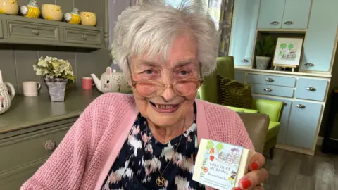 Cathy Killick/BBC An elderly woman with short grey hair smiles at the camera as she holds up a bar of soap. She has circular glasses, and wears a pink knitted cardigan over a navy printed top.
