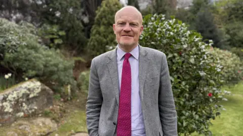 A man with a short ginger and grey bread. He slightly smiling. He is wearing a grey suit jacket, a light pink shirt and a hot pink tie.