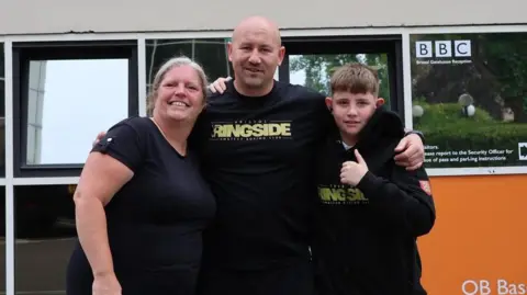 Emma, her son's boxing coach Jordan, and her son Reece are huddled for a group photograph outside a building at the BBC in Bristol in the daytime. Reece has his thumb up. They are all wearing black tops. Jordan's and Reece's tops have their boxing gym's logo on it, Bristol Ringside Amateur Boxing Club.