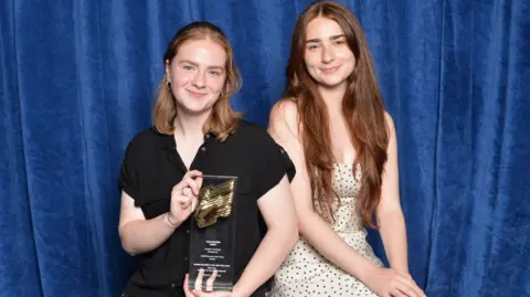 RTS/Richard Kendal Two young women stand in front of a blue velvet curtain. Niamh has short brown hair and is wearing a black, short-sleeved top. She is holding a glass award up. Amy has long brown hair and is wearing a strapless white dress with black polka dots on it. 