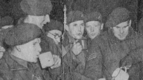 A grainy black-and-white photograph from a newspaper showing paratroopers gathered in a group drinking tea. Each is wearing their beret. It shows a moment just before they got into planes to be dropped over German lines at the River Rhine in Operation Varsity, March 1945.