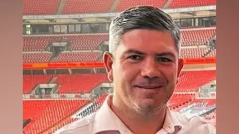 Family handout Anthony Canty is wearing a pale pink shirt inside an empty Wembley Stadium. He has short dark hair and is smiling.