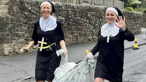 BBC/Dave Edwards Two women dressed as nuns with crosses hanging around their necks carry a bale of hay between them as they run along a road.