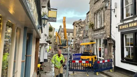 A view down Market Street in Kirkby Lonsdale. The narrow street has been cordoned off with diggers visible. Debris lies in the middle of the street in the distance and a man in hi-vis can be seen in the middle of the photo.