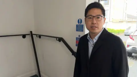 Person standing indoors next to a stairwell, wearing glasses, a dark jacket, and a light collared shirt. A handrail runs along the wall beside them, and a sign behind reads ‘Please ensure this door is closed securely on entry and exit of building.’ Through a nearby doorway or window, parked cars and buildings are visible outside