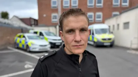 A male police officer with close cropped dark brown hair is looking at the camera with a closed mouth. He is wearing a black police uniform and there are three police vehicles in the background behind him.