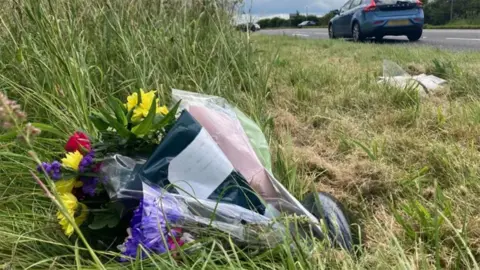A bouquet of yellow, red and purple flowers is laid on grass beside a grey road as a car drivers past. A note is attached to the bouquet.