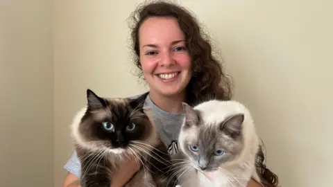 Jade Allen smiles at the camera, she has brown hair and is wearing a grey tshirt. Most of her tshirt is obscured because she is holding two cats.