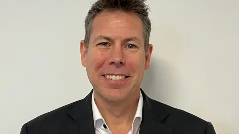 Lyle Wraxall, a man with a bright smile, he has spiky dark hair and is wearing a black jacket over a white shirt.