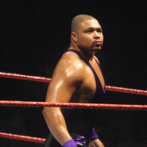 Supplied D'Lo Brown wearing a black wrestling outfit. He has purple wrist bands. He is stood in a wrestling ring.